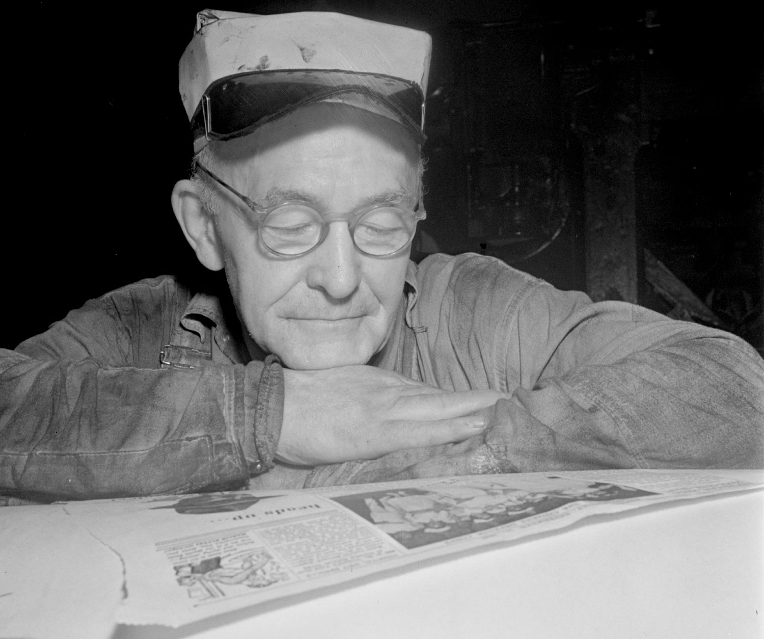 A pressman resting between editions at the New York Times in 1942, leaning on a printed page with glasses pushed up on his cap